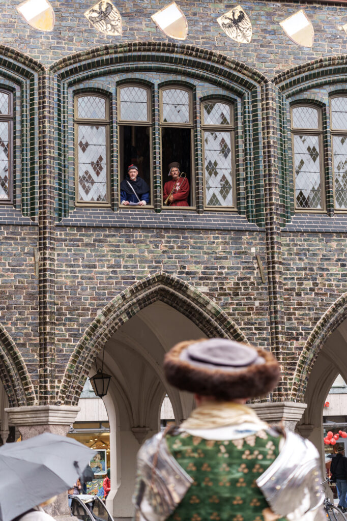 Mitglieder des Hansevolks sprechen aus einem geöffneten Fenster des Lübecker Rathauses zu den Menschen auf dem Marktplatz.